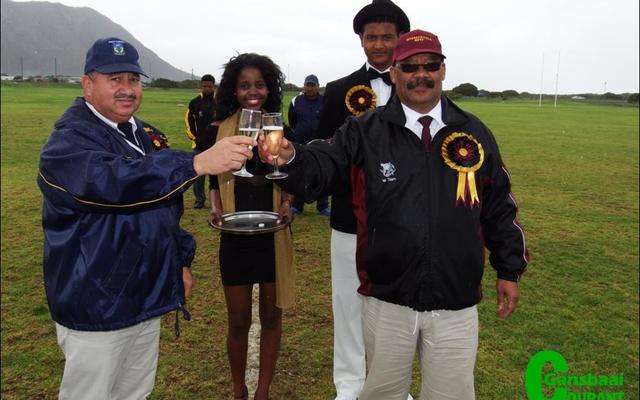 Die twee skoolhoofde, mnr Wilfred Taylor (links) van Hoërskool Eben Dönges en mnr Tommy Wilson van Gansbaai Academia klink ‘n glasie sjampanje voor die hoofwedstryd. Agter hulle is die sjampanje-nooi, Anelisa Xhego en haar begeleier, Ralph-Vino Richter.